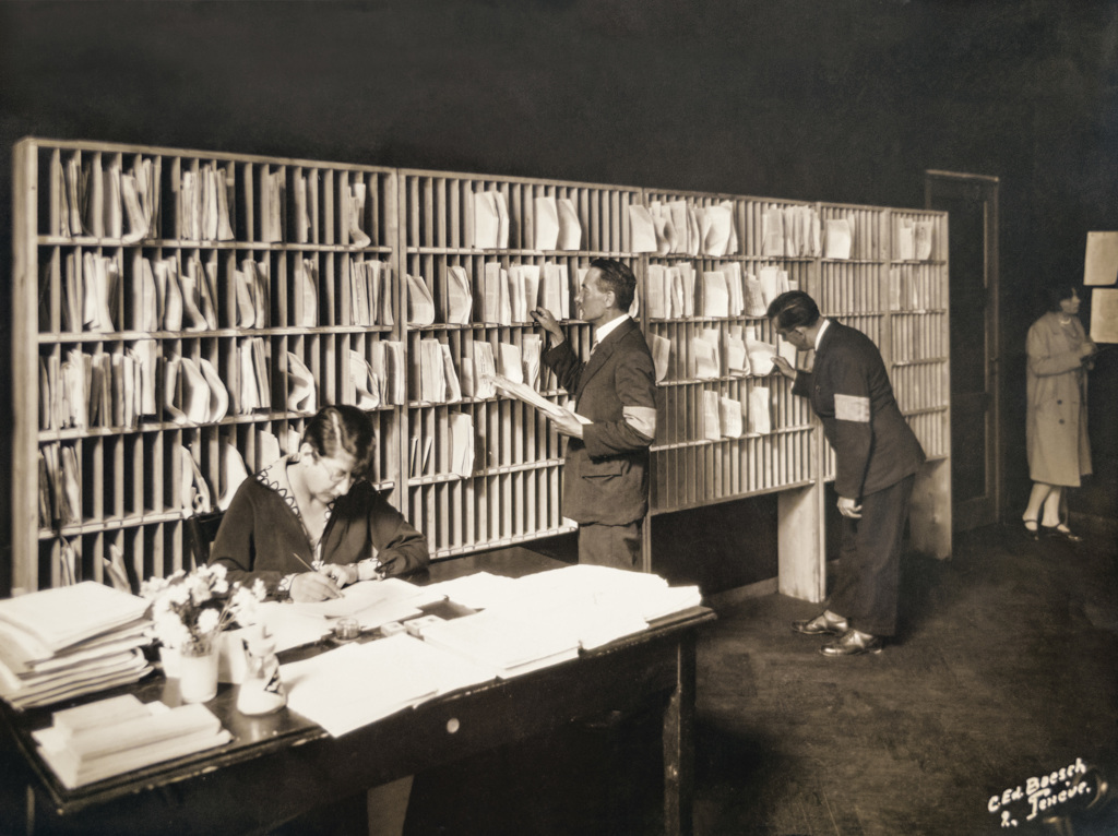 Photo en noir et blanc, service de tri du courrier - une femme assise écrivant à un bureau et deux hommes debout regardant le courrier qui est organisé dans les différents casiers contre le mur.
